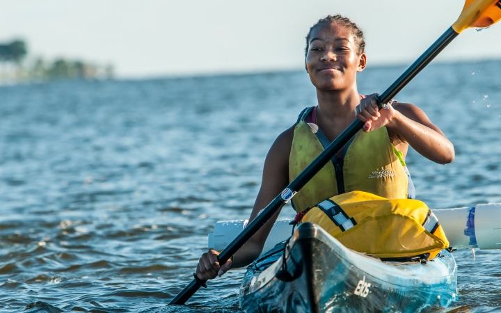 A young person smiles confidently as they paddle a kayak on very blue water.